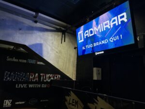 A nightclub stage with illuminated branding and a large DOOH screen displaying a promotional message, highlighting dooh screen quality in a dark indoor environment.